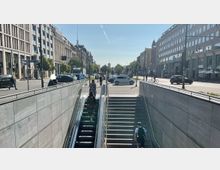 Straßenansicht mit breiter Treppe und Rolltreppe, die aus einer U-Bahn-Station führen, flankiert von modernen Bürogebäuden aus Stein und Glas. Im Hintergrund verlaufen Straßen mit Autos, Fußgängern und Radfahrern sowie vereinzelte Bäume.