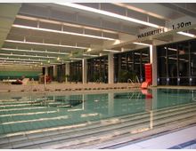 Hallenbad mit einem großen Becken, einer roten Rutsche am Beckenrand und einer Beschilderung der Wassertiefe zwischen 1,30 m und 2,20 m; im Hintergrund sind große Fenster und grüne Pflanzen zu sehen.