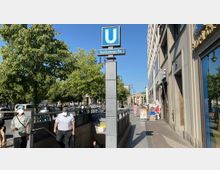 Straßenaufnahme mit U-Bahn-Schild der Station „Brandenburger Tor“ in Berlin; im Vordergrund der Treppeneingang zur U-Bahn, flankiert von Bäumen auf der linken Seite und Gebäuden mit Geschäften auf der rechten Seite. Im Hintergrund ist das Brandenburger Tor zu sehen.