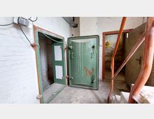 Ansicht eines Kellerraums mit weiß gestrichenen Ziegelwänden und einer grünen, alten Metalltür. Im Vordergrund sind rostfarbene Metallgeländer zu sehen, und im Hintergrund führt eine offene Tür in einen weiteren Raum.