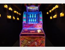 Ein bunt beleuchteter Jahrmarkt-Spielautomat mit einem auffälligen, grafischen Clown-Design steht in einem dunklen Raum, der von elektrischen Kerzen an den Wänden beleuchtet wird.