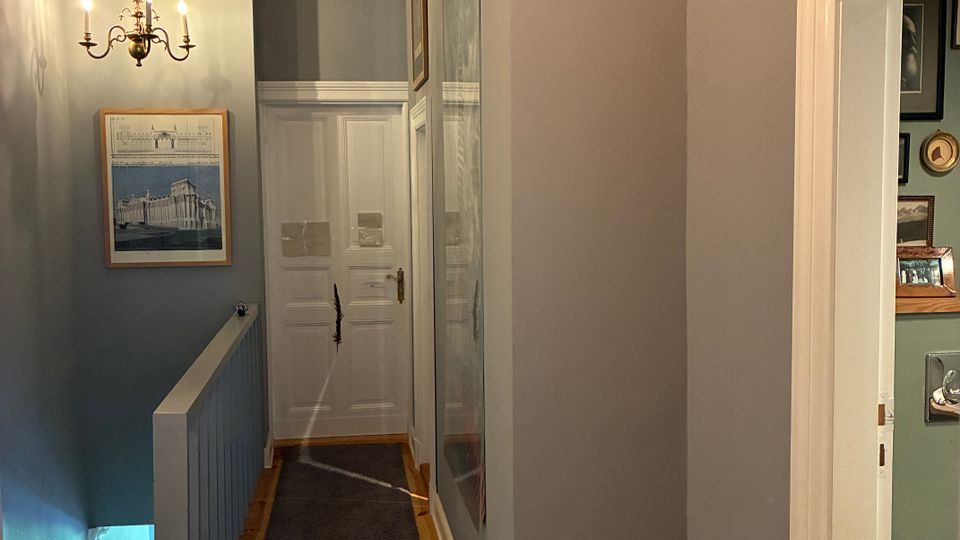 Schmaler Flur im Obergeschoss mit grauen Wänden, Holzfußboden und grauem Läufer, an dessen Ende eine weiße Tür steht. Links führt ein Treppenabgang mit Geländer nach unten, darüber hängt ein Kronleuchter; an den Wänden befinden sich gerahmte Bilder, rechts ist eine offene Tür zu einem kleinen Bad zu sehen.
