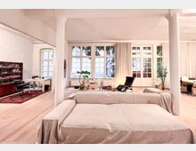 Helles Wohnzimmer mit großen, verglasten Fenstern und weißen Vorhängen, Holzboden und schlichten Möbeln, darunter ein überzogener Sofa, ein Sessel und ein Schreibtisch im Hintergrund. Die Raumgestaltung umfasst minimalistische Pflanzen und eine Bücherwand an der linken Seite.