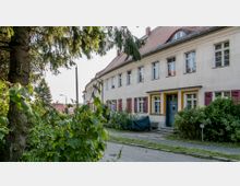 Wohngebäude mit heller Fassade und rotem Satteldach; die Fenster sind symmetrisch angeordnet und mit roten Fensterläden versehen. Im Vordergrund befinden sich dichtes Grün und ein Gehweg, der am Haus entlangführt.