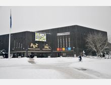 Ein verschneites Kultur- und Bildungszentrum mit moderner, dunkler Fassade und großen Glasfenstern. Im Vordergrund sind Schneebedeckung, ein Fahrradfahrer und einige Bäume zu sehen.