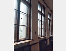 Reihe von hohen, rechteckigen Holzrahmenfenstern mit Glas, die Tageslicht in einen Innenraum lassen; darunter befinden sich Heizkörper und schmale Fensterbänke. Der Raum wirkt schlicht, mit weißen Wänden und minimaler Einrichtung.