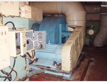 Industrieanlage mit einem großen, blau lackierten Elektromotor, umgeben von Rohren und Schutzverkleidungen. Im Vordergrund befinden sich offene Steuerkästen mit sichtbaren Kabeln und Schaltkreisen.