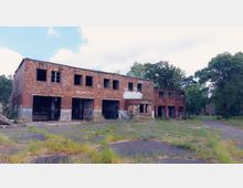 Ehemaliges Backsteingebäude mit zwei Etagen, verfallen und teilweise mit Graffiti bedeckt; Fenster und Türen fehlen, und das Gelände ist von Asphalt mit überwucherndem Gras umgeben. Im Hintergrund sind Bäume und weitere verlassene Gebäude erkennbar.