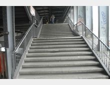 Breite Treppe aus Beton unter einer Brückenkonstruktion mit Metallgeländern auf beiden Seiten; links und rechts sind Stahlträger und Wände mit Graffiti zu sehen. Im Hintergrund gehen zwei Personen die Treppe hinauf.