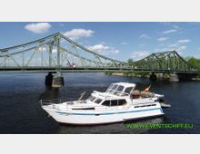 Eine grüne Stahlbrücke mit Bögen überspannt einen Fluss, darunter fährt ein weißes Motorboot mit offener Überdachung und einer deutschen Flagge am Heck. Im Hintergrund sind Bäume und ein blauer Himmel mit weißen Wolken sichtbar.
