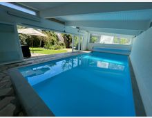 Hallenbad mit blauem Wasser und Blick nach draußen auf einen grünen Garten mit Sonnenschirm und Bäumen. Große Fensterfronten lassen viel Tageslicht in den Raum.