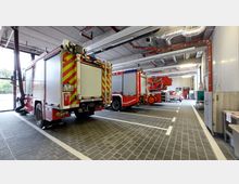 Innenansicht einer Feuerwehrgarage mit geparkten Feuerwehrfahrzeugen. Der Raum zeigt eine helle, industrielle Ausstattung mit grauen Bodenfliesen, sichtbaren Rohren an der Decke und einem offenen Tor im Hintergrund.