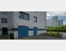 Mehrstöckiges Wohngebäude mit grauer Fassade und blauen Türen im Vordergrund, umgeben von einem gepflasterten Weg und einer kleinen Grünfläche; im Hintergrund sind weitere Wohnblocks und dichte Bäume sichtbar.