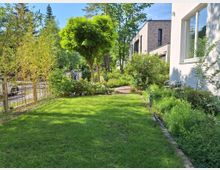 Ein gepflegter Garten mit einer Rasenfläche, umgeben von Gehölzen, Bambuspflanzen und einem kleinen Baum. Im Hintergrund sind moderne Gebäude mit Klinkerfassaden und ein Gartenweg zu sehen; rechts befindet sich die weiße Fassade eines Hauses mit Fenstern.