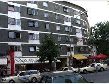 Mehrstöckiges Wohn- und Geschäftsgebäude mit grauer und weißer Fassade, zahlreichen rechteckigen Fenstern und Balkonen. Im Erdgeschoss befinden sich Geschäfte und Cafés mit gelben und roten Markisen; davor stehen geparkte Autos und ein Baum.