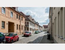 Pflasterstraße in einer ruhigen Wohngegend mit zweistöckigen Häusern aus Putz und Satteldächern, geparkten Autos entlang der Straße und vereinzelten Straßenlaternen. Der Himmel ist teilweise bewölkt und die Umgebung wirkt sauber und gepflegt.