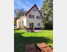 Ein Einfamilienhaus mit Spitzdach, cremefarbener Fassade und roten Fensterläden, umgeben von einem gepflegten Garten mit Rasen und Bäumen. Im Vordergrund stehen Holzliegen, und auf der Terrasse sind Blumentöpfe angeordnet.