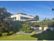 Garten mit Rasen und bepflanztem Kiesbeet, daneben eine kleine Holzterrasse mit Sitzgruppe und Sonnenschirm, umgeben von Hecken und Ziersträuchern. Im Hintergrund steht ein modernes, weißes Flachdachhaus mit großen Fenstern vor blauem Himmel.