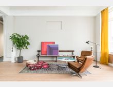 Wohnzimmer mit minimalistischem Design: Ein brauner Ledersessel steht neben einem kleinen runden Tisch, auf dem ein aufgeschlagenes Buch liegt. Im Hintergrund sind eine Holzbank mit bunten Bildern darauf, ein großer Topfpflanze, eine Stehlampe und gelbe Vorhänge vor einem Fenster zu sehen, der Boden ist mit einem gemusterten Teppich bedeckt.