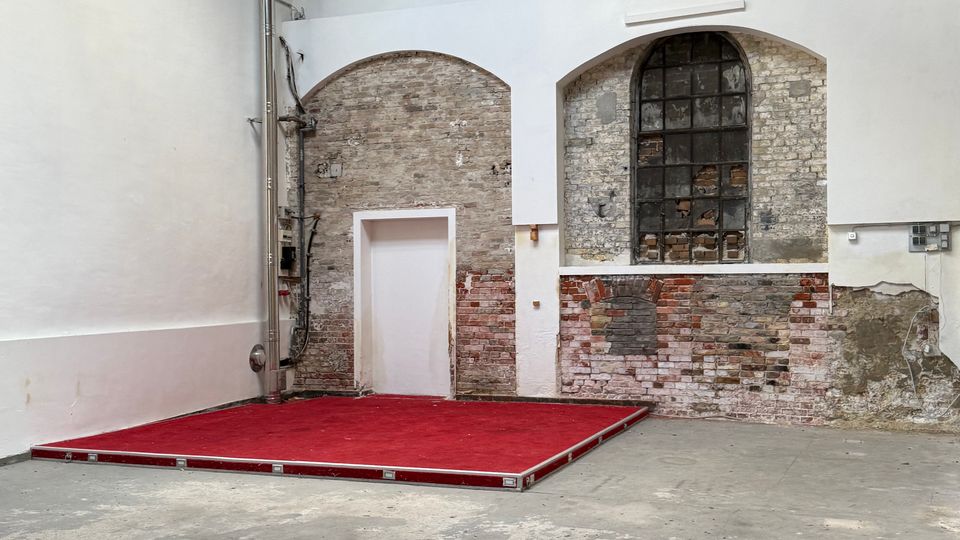 Innenraum einer ehemaligen Industriehalle mit hohen weißen Wänden und freigelegtem Ziegelmauerwerk in Rundbogen-Nischen, darunter ein kleines weißes Türportal. Rechts befindet sich ein großes, bogenförmiges Sprossenfenster; die Decke besteht aus Holz und sichtbaren Stahlträgern, am Boden steht eine niedrige Bühne mit rotem Teppich.