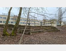 Blick auf ein modernes, mehrstöckiges Schul- oder Bürogebäude mit vielen rechteckigen Fenstern. Davor liegen terrassenförmig angelegte Betonstufen in einem kahlen, laubbedeckten Außenbereich mit mehreren Bäumen.