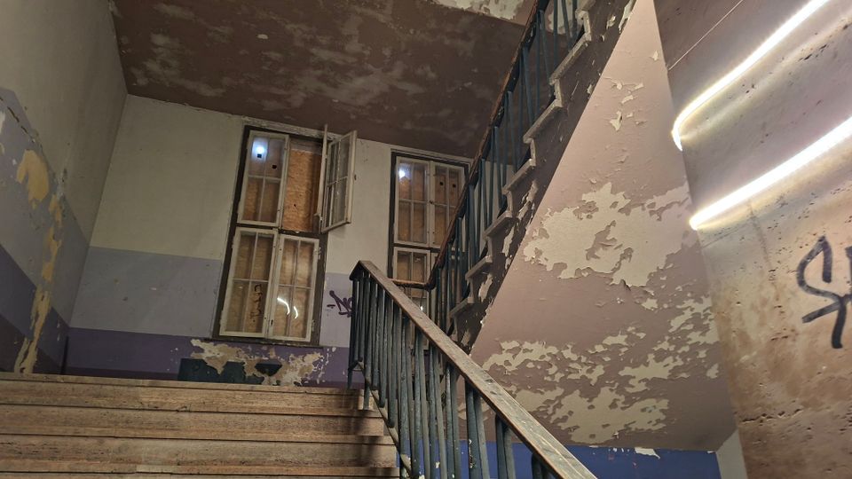 Abgenutztes Treppenhaus in einem älteren Gebäude mit Holzstufen, Metallgeländer und stark abblätternder Farbe an den Wänden. Zwei hohe Fenster mit teils geöffneten Flügeln und abgedeckten Scheiben lassen Licht herein, an den Wänden sind Graffiti zu sehen.
