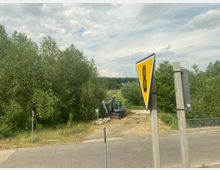 Eine ländliche Straße mit einem gelben Verkehrszeichen im Vordergrund. Im Hintergrund befindet sich ein kleiner Bagger neben einer grasbewachsenen Fläche, umgeben von Bäumen und dichter Vegetation.