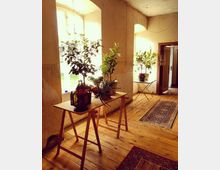 Heller Innenraum mit Holzdielenboden und einem Tisch aus Holz, auf dem Zimmerpflanzen und eine orientalische Laterne stehen. Im Hintergrund sind große Fenster mit Blick ins Freie und weitere Pflanzen auf einem Beistelltisch zu sehen.