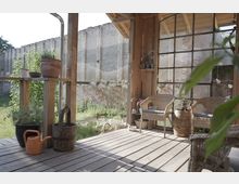 Holzveranda mit Sitzecke, bestehend aus zwei Korbstühlen und einem Fass-ähnlichen Tisch, umgeben von Holzwänden und einer Fensterfront mit Metallrahmen. Im Hintergrund eine alte Backsteinmauer und grüne Vegetation.