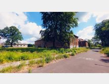 Backstein-Gebäude in einem verlassenen Zustand, umgeben von überwucherten Grünflächen und kleinen Bäumen; eine asphaltierte Straße führt am Gebäude entlang. Einige Fassaden sind mit Graffiti bedeckt, und der Himmel ist leicht bewölkt.