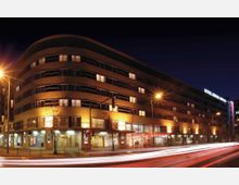 Nachtszene eines modernen Hotels mit abgerundeter Fassadenarchitektur und mehreren Fensterreihen, beleuchtet mit warmem Licht. Im Vordergrund sind Straßenlaternen, Bäume und Lichtspuren des Verkehrs sichtbar.