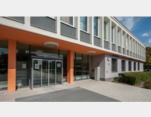 Moderne Fassade eines Amtsgerichtsgebäudes mit weitläufiger Glasfront im Eingangsbereich, orangefarbenen Säulen, weißen Betonrahmungen und schmalen Fenstern. Der Vorplatz ist mit Pflastersteinen bedeckt und von niedrigen Hecken gesäumt.