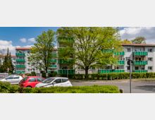 Mehrstöckige Wohngebäude mit weißen Fassaden und grünen Balkonverkleidungen, umgeben von Bäumen und Hecken. Im Vordergrund befindet sich eine Straße mit geparkten Autos und Verkehrszeichen.