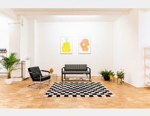 Minimalistisch eingerichtetes Wohnzimmer mit Holzboden, einer schwarz-weiß gestreiften Bank, einem schwarzen Ledersessel, einem gläsernen Couchtisch und einem schwarz-weiß karierten Teppich. An der weißen Wand hängen zwei bunte, abstrakte Kunstwerke; Pflanzen und Regale ergänzen die Dekoration.