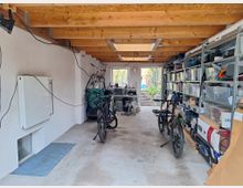 Garage mit offener Holzbalkendecke, einer Werkbank im Hintergrund und Regalen voller Werkzeug und Kisten auf der rechten Seite. Zwei Fahrräder und diverses Zubehör befinden sich im Vordergrund, und durch eine Glastür ist ein Garten erkennbar.