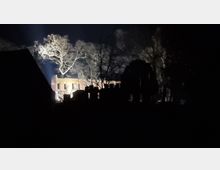 Nachtszene einer beleuchteten Backsteinruine, umgeben von Bäumen. Im Vordergrund dunkle Silhouetten von Strukturen und Ästen, während ein großer beleuchteter Baum deutlich hervorsticht.