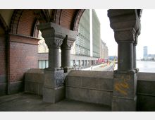 Blick von einem gemauerten Durchgang mit verzierten Steinsäulen und Rundbögen aus rotem Backstein auf moderne Gebäude mit Glasfassaden und die Uferlinie eines Flusses. Im Hintergrund sind ein breiter Gehweg und eine städtische Umgebung sichtbar.