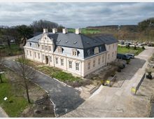 Luftaufnahme eines historischen Herrenhauses mit cremefarbener Fassade und dunkelgrauem Mansarddach, umgeben von einem parkähnlichen Gelände mit Bäumen und Wiesen. Ein geschotterter Weg führt zum Gebäude, während mehrere Fahrzeuge auf dem Parkplatz daneben stehen.