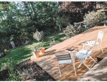 Terrasse mit Holzboden, darauf drei weiße Gartenstühle um einen kleinen Tisch. Im Hintergrund Gartenbereich mit grüner Wiese, Topfpflanzen und Bäumen.