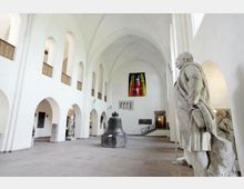 Innenraum einer historischen Kirche oder eines Museums mit gotischen Gewölbedecken, weißen Wänden und Rundbögen. Im Vordergrund steht eine große Marmorskulptur, in der Mitte eine massive Glocke, und an der Rückwand hängt ein farbenfrohes Wandgemälde.