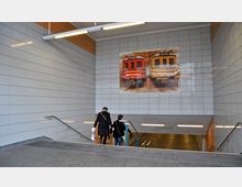 Treppenabgang zu einer U-Bahn-Station mit weißen Wandfliesen und moderner Beleuchtung. An der Wand hängt ein großes Poster mit Abbildungen von zwei historischen U-Bahn-Wagen, gekennzeichnet mit „Alexanderplatz“ und „Friedrichstraße“.