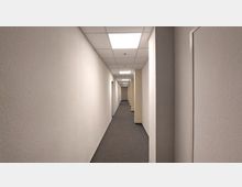 Ein langer Flur mit weißen Wänden, grauem Teppichboden und einer Decke mit rechteckigen, eingebauten LED-Leuchten; seitlich befinden sich mehrere Türen.