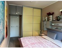 Schmales, modern eingerichtetes Schlafzimmer mit Doppelbett und rosa Tagesdecke, seitlichen Regalen und Wanddekorationen. Am Ende des Raums stehen eine weiße Tür sowie ein großer Schiebetür-Kleiderschrank mit hellgelben Paneelen und grauem Rahmen.