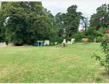 Eine weitläufige grüne Wiese mit einem kleinen, grünen Geräteschuppen im Hintergrund, umgeben von hohen Bäumen und einer weiß verputzten Mauer, teilweise bewachsen mit Efeu; rechts im Vordergrund blühende Rosensträucher.