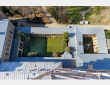 Luftaufnahme eines quadratischen Klosterinnenhofs mit Rasenfläche, umgeben von überdachten Arkadengängen mit Rundbögen. Die Gebäude haben blaue Metalldächer, und ein kleiner Brunnen mit Statue ist im rechten Bereich des Hofs zu sehen; umliegend befinden sich Gehwege und Bäume.