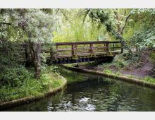 Eine kleine, hölzerne Fußgängerbrücke überquert einen schmalen Fluss oder Kanal, umgeben von dichtem, grünem Laub und überhängenden Bäumen. Die Ufer sind mit Gras und niedriger Vegetation bewachsen, und der Kanal ist von einer schmalen Ziegelmauer eingefasst.