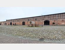 Großer gepflasterter Hof mit einem langgestreckten Backsteingebäude im Fachwerkstil im Hintergrund. Das Gebäude besitzt große Holztore und kleine Fenster, während im Vordergrund ein kleiner gemauerter Brunnen steht.