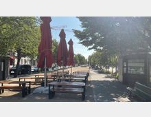 Straßencafé mit Holzsitzbänken und geschlossenen roten Sonnenschirmen auf einem gepflasterten Weg, umgeben von Bäumen. Im Hintergrund sind Menschen, parkende Autos und ein Verkaufsstand mit Speiseangeboten erkennbar.