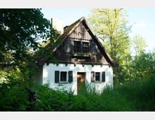 Kleines Fachwerkhaus inmitten einer grünen, bewaldeten Landschaft, mit weiß gestrichenen Wänden, dunklen Holzfensterläden und einem steilen Dach mit braunen Ziegeln. Vor dem Haus wächst hohe Vegetation, umgeben von Bäumen und Sträuchern.