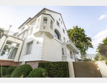 Weiße zweistöckige Villa mit roten Dachziegeln und dekorativer Fassade, großen Fenstern und einem Erker. Umgeben von gepflegten Büschen und Bäumen, mit einem metallischen Tor und klarem Himmel im Hintergrund.